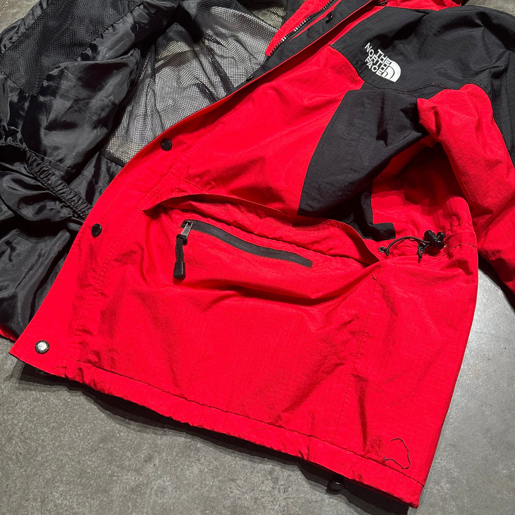 A red and black The North Face jacket laid flat on a gray concrete floor, showing the outer shell, inner zip pocket, and part of the black hood.